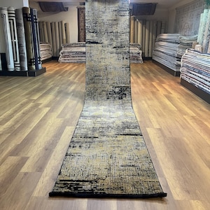 May include: A black and gold runner rug with a geometric pattern. The rug is rolled up and standing on a wooden floor.