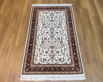May include: Rectangular rug with a cream-colored center featuring a floral pattern in shades of red, gold, and brown. The border is a deep burgundy with intricate designs. White fringe trims the edges.