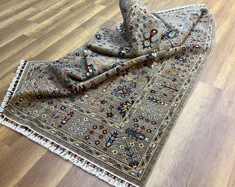 Könnte beinhalten: Ein beiger Teppich mit Blumenmuster und einem weißen Fransenrand. Der Teppich ist in der Mitte gefaltet und liegt auf einem Holzboden.