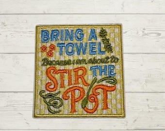 May include: A square embroidered patch featuring a yellow and white gingham background. The text reads BRING A TOWEL in blue, followed by Because I am about to in black, and STIR THE POT in orange. Decorative green vines and small orange flowers are included.