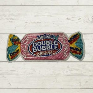 May include: An embroidered patch shaped like a piece of wrapped candy. The center is pink with the text DOUBLE BUBBLE ORIGINAL in blue and red. The twisted ends are yellow and teal with the text DOUBLE BUBBLE embroidered in small letters.
