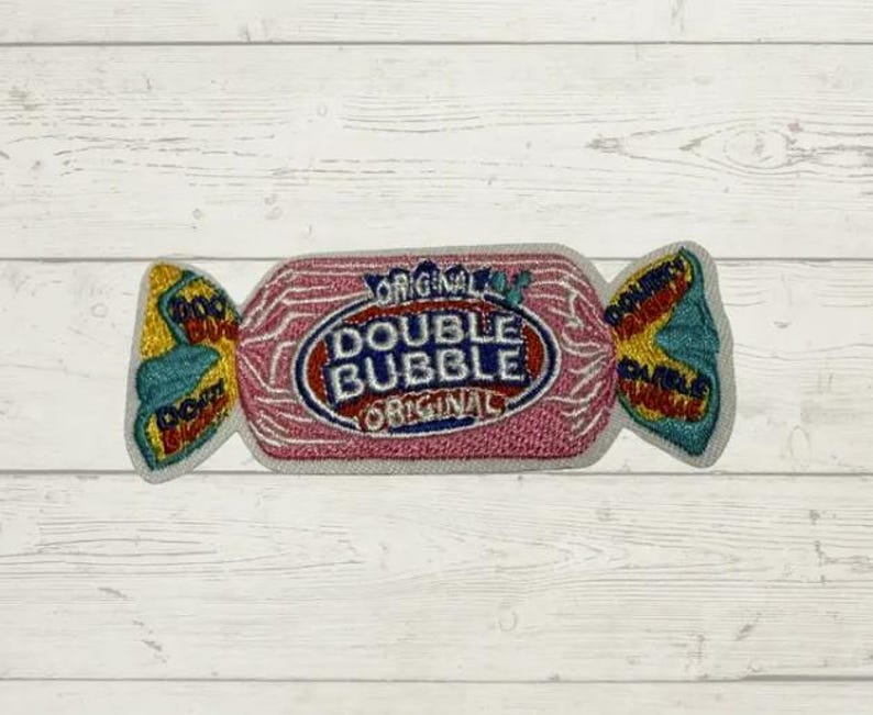 May include: An embroidered patch shaped like a piece of wrapped candy. The center is pink with the text DOUBLE BUBBLE ORIGINAL in blue and red. The twisted ends are yellow and teal with the text DOUBLE BUBBLE embroidered in small letters.