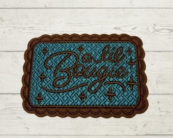 May include: A rectangular embroidered patch with a scalloped brown border. The center features a teal background with a woven texture, dark brown embroidered text reading "a lil Bougie," and small dark brown four-pointed star accents.