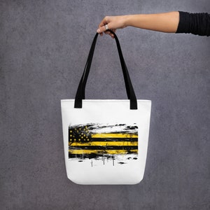 Puede incluir: Una bolsa de tela blanca con asas negras y un diseño de bandera estadounidense negra y amarilla. La bandera está desgastada y tiene un efecto grunge.