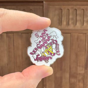 Puede incluir: Un pin de esmalte blanco con una ilustración detallada de una estructura de proteína en tonos magenta, amarillo y blanco. El pin se sostiene entre los dedos sobre un fondo de madera.