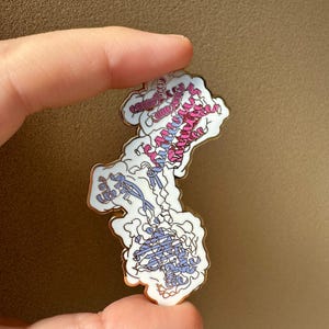 Puede incluir: Pin de esmalte detallado con un diseño de estructura molecular en tonos rosa, azul y blanco, con un contorno dorado. El pin se sostiene sobre un fondo neutro, mostrando sus intrincados detalles y su temática científica.