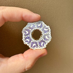 May include: A circular enamel pin with a gold border, showcasing a repeating floral pattern in purple and white. The pin is held by fingers against a plain background.