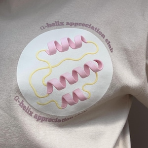 以下が含まれることがあります： ベージュのクルーネックTシャツ。円形のグラフィックには、ピンクのアルファヘリックスのデザインと黄色の線が描かれています。「α-helix appreciation club」の文字が薄紫でプリントされています。柔らかい素材のシャツです。