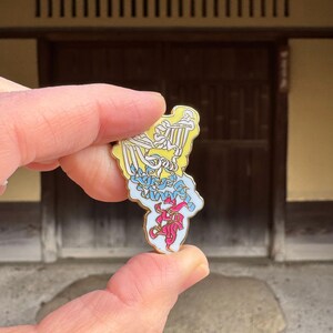 以下が含まれることがあります： 黄色、白、青、ピンクのアクセントが付いた、スタイリッシュな鳥のデザインが施された金色のエナメルピン。ピンは手で持たれています。