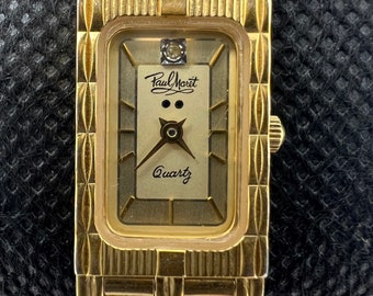 May include: A gold-tone wristwatch with a rectangular face. The watch face has a white background with black Roman numeral hour markers and a small diamond at the 12 o'clock position. The watch face also features the text "Paul Morlet" and "Quartz".