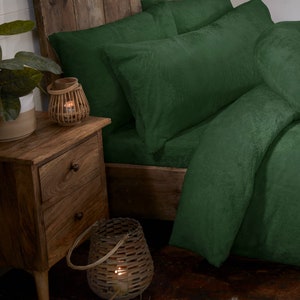 May include: A green plush bedding set with a pillow and a bolster pillow. The bedding is on a wooden bed frame with a wooden bedside table next to it. There are two wicker candle holders on the bedside table.