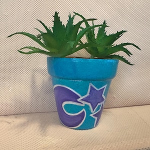 Könnte beinhalten: Ein handbemalter teal- und blauer Blumentopf mit einem lila Stern- und Halbmond-Design. Der Topf enthält eine grüne Sukkulentenpflanze. Der Topf ist ein dekorativer Artikel für die Inneneinrichtung.