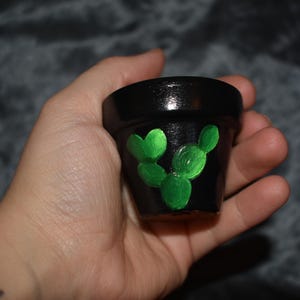 May include: A small, black ceramic plant pot with a hand-painted green cactus design. The pot is cylindrical and has a glossy finish. The cactus design features three green, rounded shapes. The pot is held in a hand.