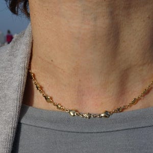Può includere: Una collana d'oro con una catena delicata e ciondoli a forma di cuore. La collana è indossata da una persona con un maglione grigio.