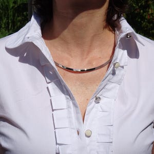 Può includere: Una collana girocollo color argento. La collana è una semplice fascia curva. La persona indossa una camicia bianca con colletto, pieghe decorative e bottoni.
