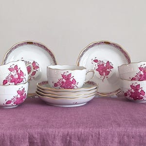 May include: A white porcelain tea set with a floral design in shades of pink and gold trim. The set includes teacups, saucers, bowls, and plates. The items are arranged on a purple tablecloth.