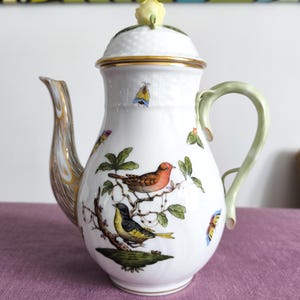 May include: White porcelain coffee pot with a lid and a curved handle. The pot is decorated with colorful birds, butterflies, and green leaves. The lid is topped with a yellow rosebud and has a gold trim.