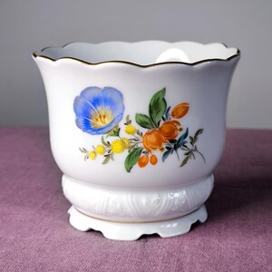 Op de afbeelding: Een witte porseleinen plantenpot met een geschulpte rand en voet, voorzien van een gouden randje. De buitenkant is versierd met een bloemmotief bestaande uit een grote blauwe bloem, kleine gele bloesems en oranje vruchtachtige elementen.