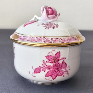 Può includere: Una zuccheriera in porcellana bianca con coperchio, con disegni floreali rosa e un bordo dorato. Il coperchio è sormontato da una rosa rosa. La ciotola ha una superficie strutturata, possibilmente vintage.