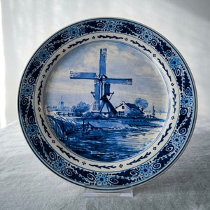 May include: A blue and white ceramic plate with a windmill scene. The plate features a traditional Dutch windmill with a blue and white design. The plate is decorated with a blue and white border.