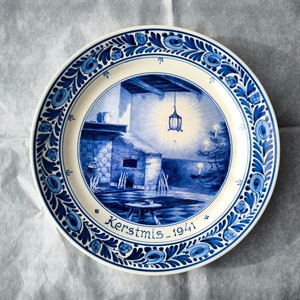 Può includere: Un piatto in ceramica blu e bianco con una scena natalizia festosa. Il piatto presenta un design dipinto in blu e bianco di un camino, un albero di Natale e una lanterna. Il testo "Kerstmis 1941" è scritto sul piatto.