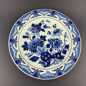 May include: A white ceramic plate with a blue and white floral design. The plate features a traditional Dutch Delftware pattern with intricate floral motifs and a blue border.