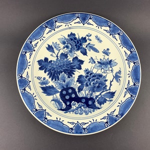 May include: A white ceramic plate with a blue and white floral design. The plate features a stylised floral pattern with blue leaves and flowers on a white background. The rim of the plate is decorated with a blue and white geometric pattern.