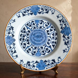 Op de afbeelding: Een ronde keramische bord met een witte basis en ingewikkelde blauwe bloemmotieven. Het midden toont een grote gestileerde bloem omringd door krullende ranken, terwijl de brede rand is versierd met herhalende blauwe bloesems en bladeren.