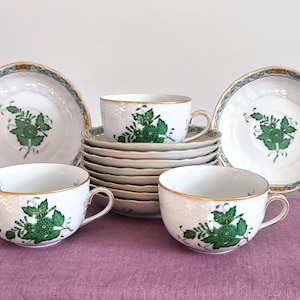 Puede incluir: Un juego de té de porcelana blanca con un diseño floral verde. El juego incluye tazas, platillos y platos. Los bordes están adornados con oro y un borde verde y dorado. El juego está dispuesto sobre un mantel púrpura.