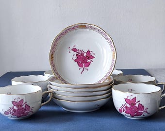 May include: A set of white porcelain teacups and saucers with a floral design in fuchsia and gold trim. The teacups and saucers are arranged on a blue surface. The set includes six saucers and four teacups.