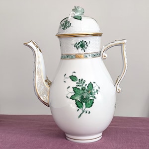 Puede incluir: Una cafetera de porcelana blanca con tapa, asa y boquilla. La cafetera está decorada con diseños florales verdes y ribetes dorados. La tapa tiene un pequeño acento floral verde. La cafetera está sobre una superficie morada.