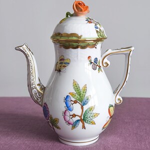 Puede incluir: Una tetera de porcelana blanca con un motivo floral y de mariposas pintado a mano en colores vibrantes. La tapa está rematada con una rosa naranja esculpida. La tetera presenta un asa curva, un pico decorativo y detalles en oro.