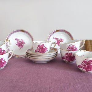 May include: A white porcelain tea set with a floral design in shades of pink and gold trim. The set includes teacups, saucers, and plates, arranged on a purple tablecloth. The teacups have a scalloped edge.