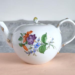 Può includere: Teiera in porcellana bianca con corpo arrotondato, con un design floreale arancione, viola e blu. La teiera ha un beccuccio curvo, un manico ad anello e finiture dorate. Un piccolo pomello con punta gialla si trova sopra il coperchio.