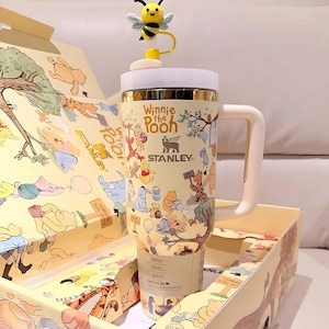 May include: A beige Stanley tumbler featuring Winnie the Pooh characters and the text "Winnie the Pooh". The tumbler has a white handle and lid, with a yellow bee-shaped topper. The tumbler is in a matching box with illustrations.