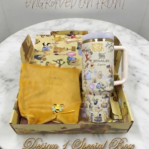May include: A special gift box featuring a Winnie the Pooh themed Stanley tumbler with a handle, a yellow pouch, and a decorative box. The tumbler has a cream background with cartoon illustrations. The box and pouch are yellow with cartoon designs. The text "Engraved on Front" is visible.