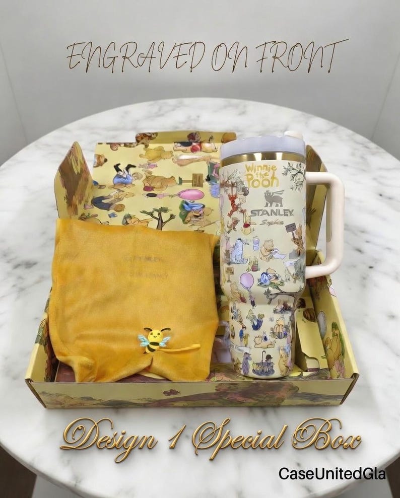 May include: A special gift box featuring a Winnie the Pooh themed Stanley tumbler with a handle, a yellow pouch, and a decorative box. The tumbler has a cream background with cartoon illustrations. The box and pouch are yellow with cartoon designs. The text "Engraved on Front" is visible.
