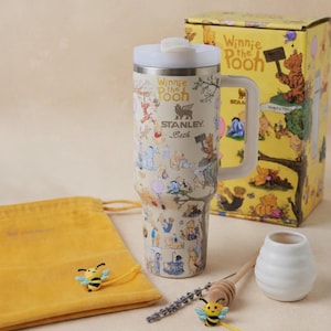 May include: A Winnie the Pooh themed gift set featuring a stainless steel tumbler with a handle, a yellow gift box, a yellow drawstring bag, and decorative bee accents. The tumbler is covered in cartoon illustrations and has a white lid. The gift set includes a honey dipper and a small white vase.