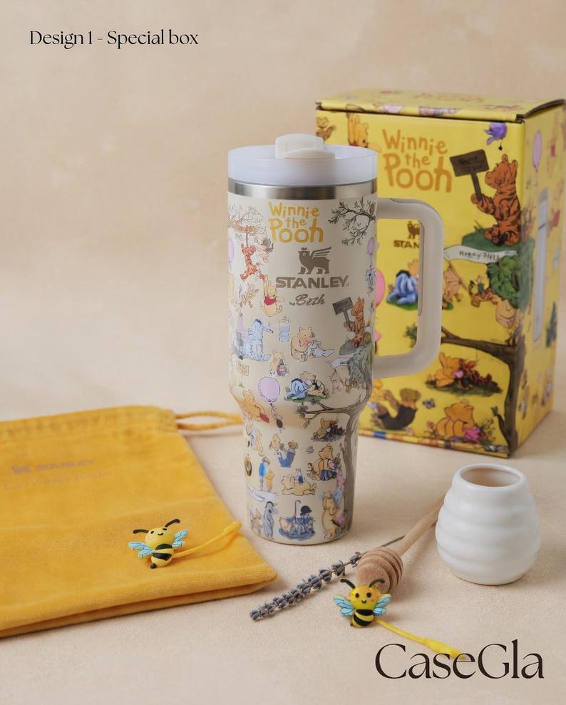 May include: A Winnie the Pooh themed gift set featuring a stainless steel tumbler with a handle, a yellow gift box, a yellow drawstring bag, and decorative bee accents. The tumbler is covered in cartoon illustrations and has a white lid. The gift set includes a honey dipper and a small white vase.