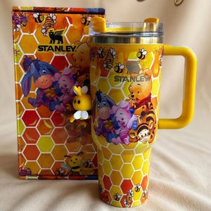 May include: Yellow Stanley tumbler and matching box featuring a honeycomb pattern and images of Winnie the Pooh and friends. The tumbler has a yellow handle and lid, with the Stanley logo visible. The box reads "Design 2 - Special box".