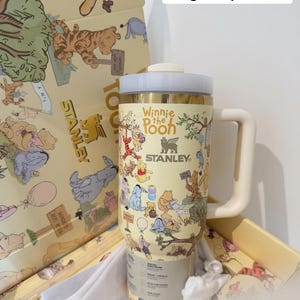 May include: A yellow Stanley tumbler featuring Winnie the Pooh characters. The tumbler has a cream-colored handle and lid, with the text "Winnie the Pooh" and "Stanley" printed on it. The tumbler is in a matching box with character illustrations.