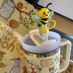 May include: A Winnie the Pooh themed Stanley mug with a yellow and black bee straw topper. The mug has a white handle and a lid. The mug is decorated with illustrations of Winnie the Pooh characters.