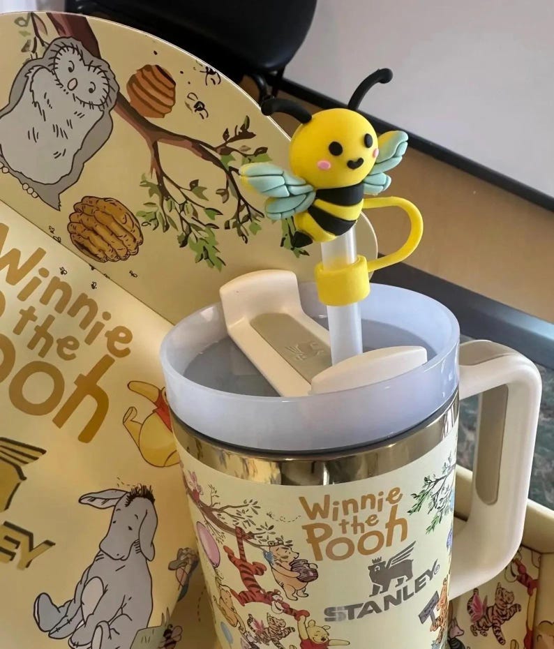 May include: A Winnie the Pooh themed Stanley mug with a yellow and black bee straw topper. The mug has a white handle and a lid. The mug is decorated with illustrations of Winnie the Pooh characters.