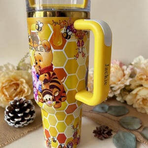 May include: Yellow tumbler with a Winnie the Pooh design. The tumbler features a honeycomb pattern, bees, and images of Winnie the Pooh, Tigger, and Piglet. The handle is yellow with a white accent and the name "Jackie" printed on it. The text "Design 2 - Special box" is visible.