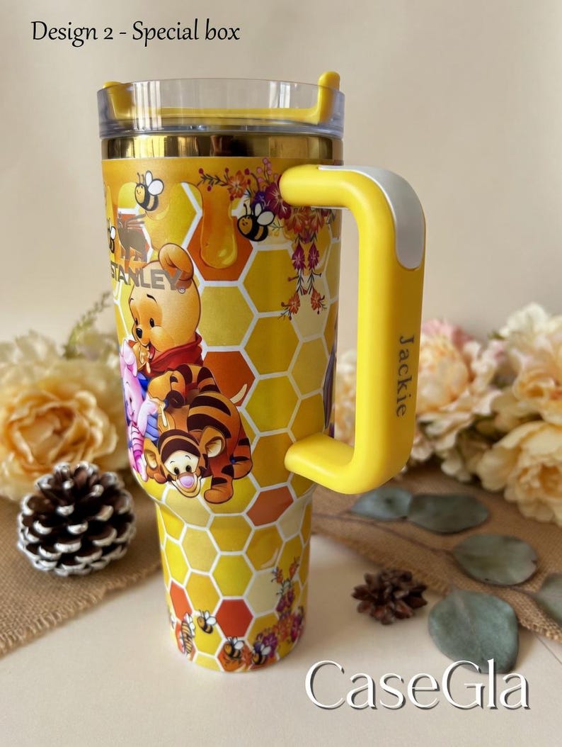 May include: Yellow tumbler with a Winnie the Pooh design. The tumbler features a honeycomb pattern, bees, and images of Winnie the Pooh, Tigger, and Piglet. The handle is yellow with a white accent and the name "Jackie" printed on it. The text "Design 2 - Special box" is visible.