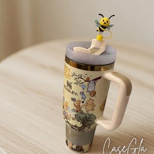 May include: A beige Stanley travel mug featuring Winnie the Pooh characters and a bee-shaped straw topper. The mug has a light beige handle and a gold rim. The text "Case Gla" and copyright information are visible.