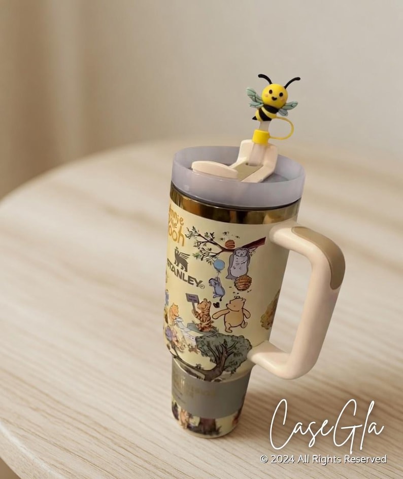 May include: A beige Stanley travel mug featuring Winnie the Pooh characters and a bee-shaped straw topper. The mug has a light beige handle and a gold rim. The text "Case Gla" and copyright information are visible.