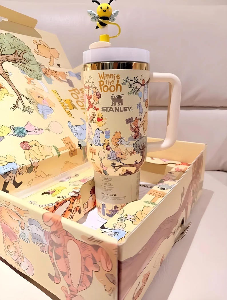 May include: A beige Stanley tumbler featuring Winnie the Pooh characters and the text "Winnie the Pooh". The tumbler has a white handle and lid, with a yellow bee-shaped topper. The tumbler is in a matching box with illustrations.