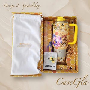May include: A gift box featuring a yellow and gold Stanley tumbler with Winnie the Pooh characters. The box includes a white drawstring bag, a straw, and a bee-shaped accessory. The box has a honeycomb pattern.