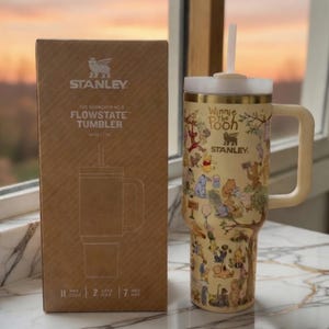 May include: A beige Stanley Flowstate tumbler with a Winnie the Pooh design, a white straw, and a handle. The tumbler is next to its cardboard box. The box has the Stanley logo and product information. The tumbler holds 40 oz.